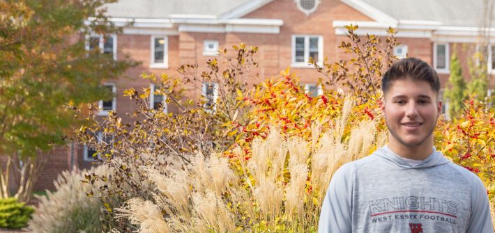 Christopher standing in front of Chestnut Hall. There is green and white shrubbery around him. Trees with green and orange leaves.