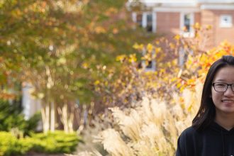 Lili poses in front of yellow and orange bushes. There is a brick building in the background and a tree with green leaves.