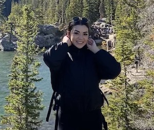 Erica pulls her hair back, while standing in the woods with a lake behind her. 