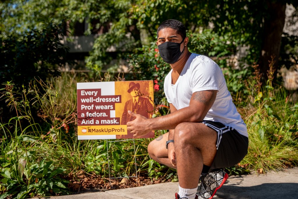 Trevor kneeling down in front of a sign with a mask on.