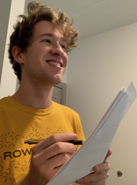 Nick wears a yellow Rowan shirt, smiles off to the distance while writing something. 