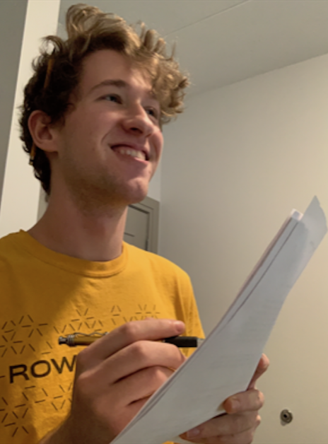Nick wears a yellow Rowan shirt, smiles off to the distance while writing something.