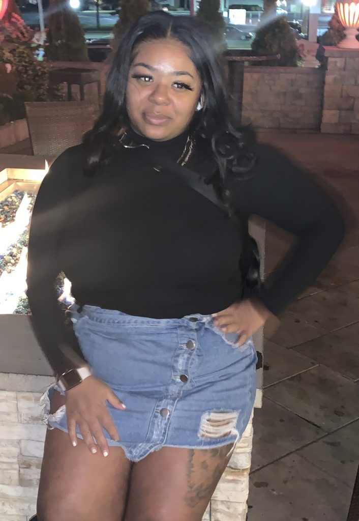 Tamora stands outdoors in a denim skirt, hand on hip.