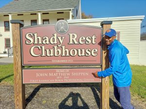 Brad posing with the Shady Rest Clubhouse sign and pointing to the name 'John Matthe Shippen'.