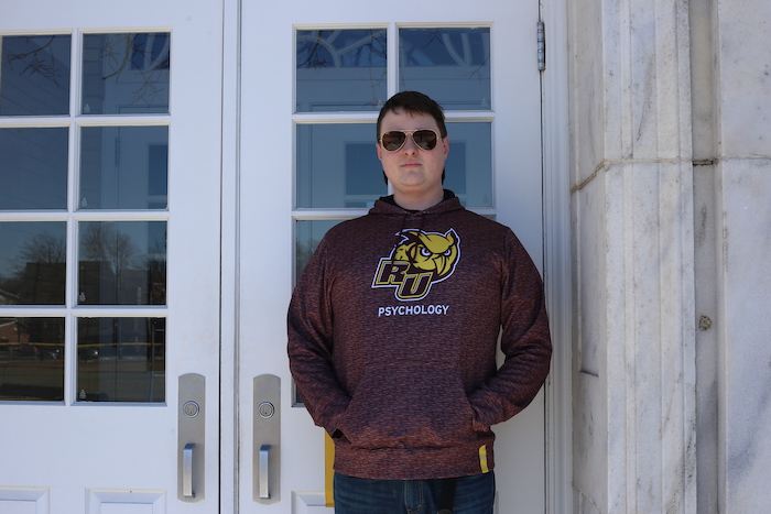 John standing against the door of Bunce while wearing sunglasses and a Rowan Psychology sweatshirt. 