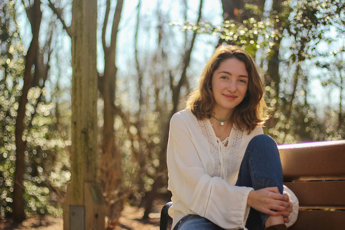 Sarah smiling and sitting on a bench outside on the campus trail.