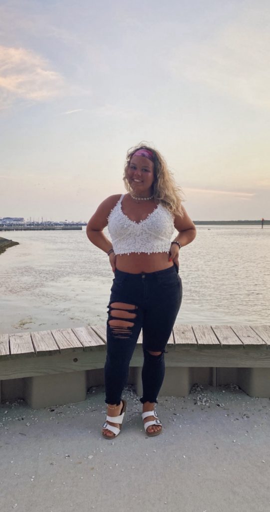 Gianna posing near water in black jeans and a white tank top.