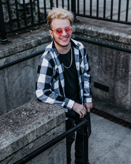 Dom smiles up at the camera by Bunce Hall, wearing black-and-white plaid and pink sunglasses.