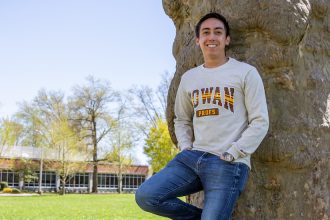 JT leans against a tree on Bunce green, wearing a Rowan sweater with hands in his pockets.