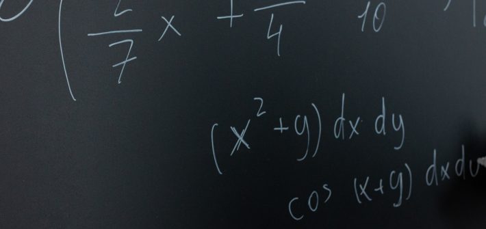 Stock image of math equations being written on a blackboard with chalk in hand.