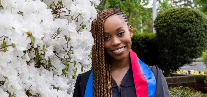Esther wears her graduation gown and stands in front of campus greenery.