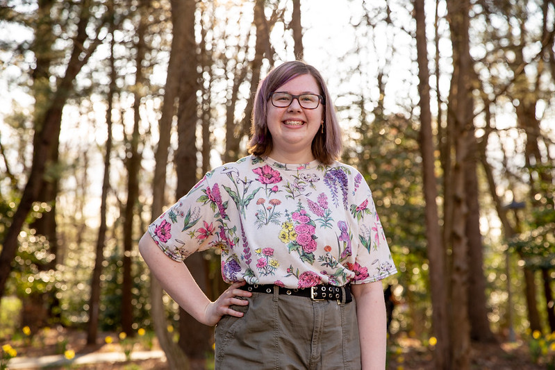 Isabel smiles, has one hand on her hip in a wooded area on campus.
