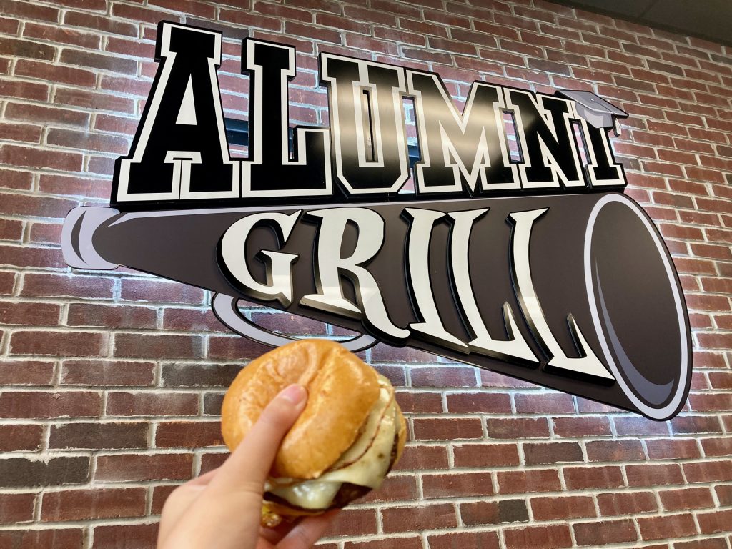 Holding up a burger to the Alumni Grill sign. 