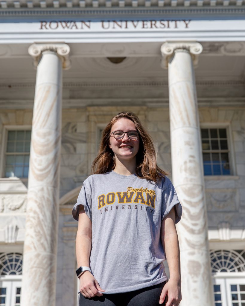 Leah wears glasses and a Rowan t-shirt smiling in front of Bunce Hall.
