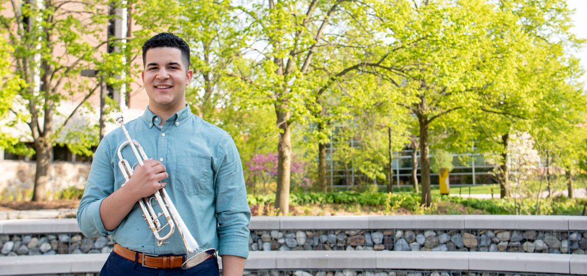 Luis stands with his trumpet outside on campus.