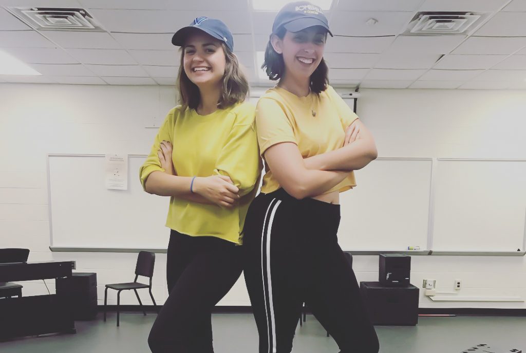 From left to right stands Angela and Molly Jo in a rehearsal studio at Rowan University. Both have their arms crossed, wearing navy blue hats and yellow shirts, with smiles on their faces.