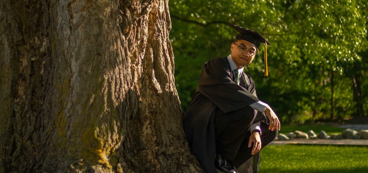 Riel wears his graduation regalia and squats by a tree.