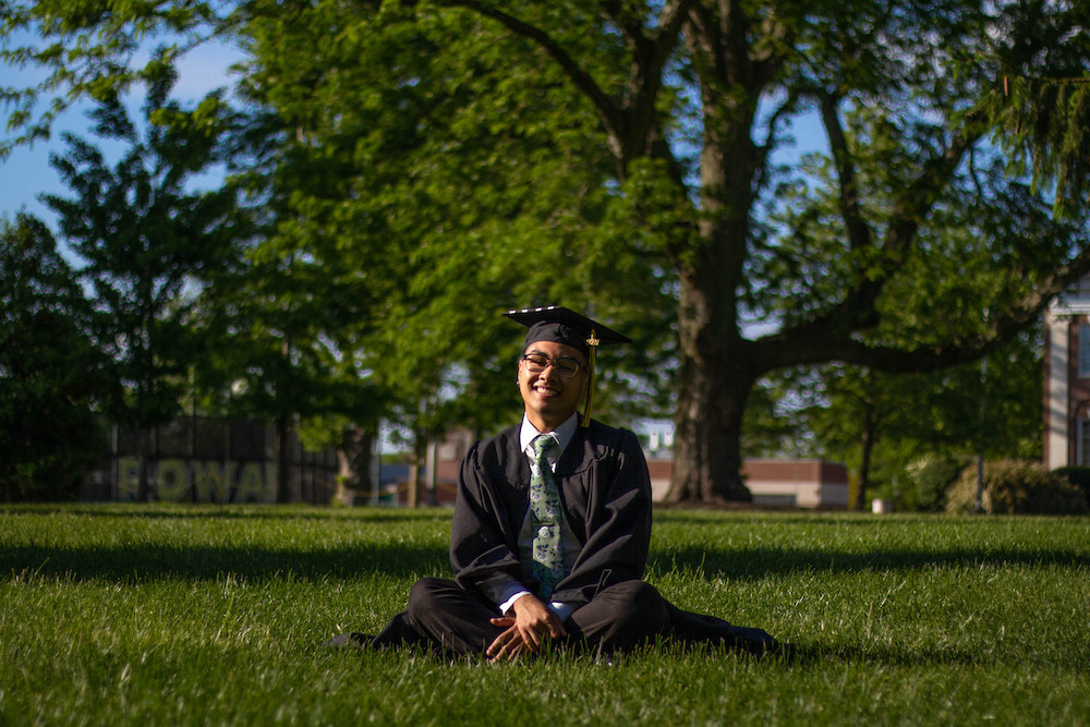 Riel sits contently in the lawn of Bunce Hall while wearing a graduation cap and gown.