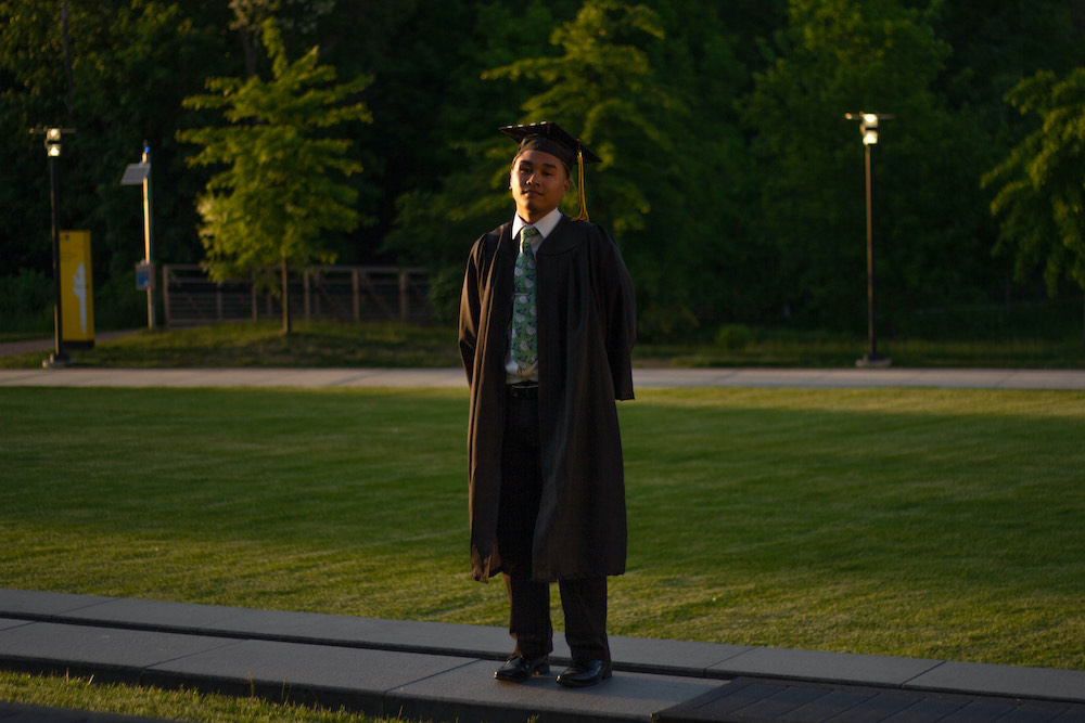 Riel stands on the lawn of Business Hall while wearing graduation regalia.