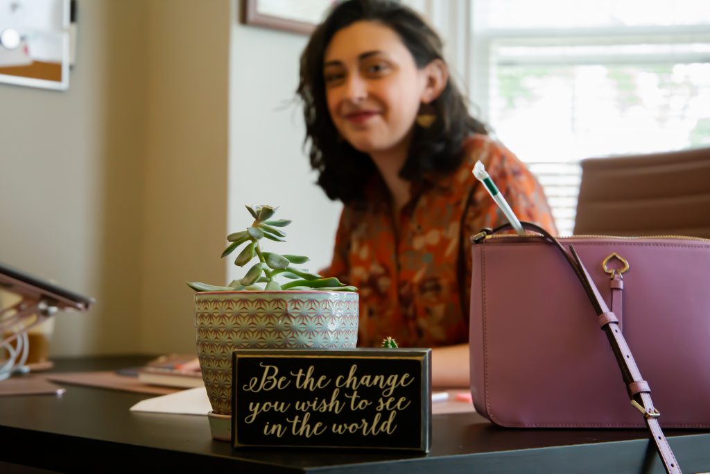 In the front is a focus on a sign on Molly Jo's desk that says "Be the change you wish to see in the world." She sits behind it out of focus with a smile on her face.