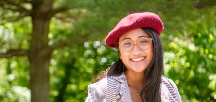 Stephanie wears glasses and a red beret smiling at the camera with a green forest in the background.