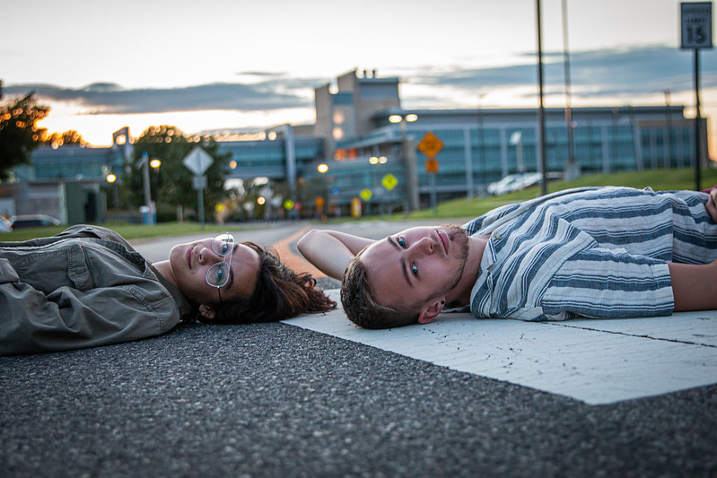 Artsy photo of two students laying down on a road, head to head, with the engineering building behind them.