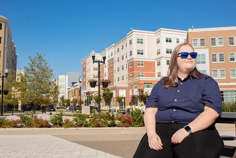 Carrie sits on a bench on Rowan Boulevard wearing sunglasses and looking away.