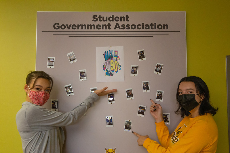 Madeline and Sarah pose in front of the SGA bulletin board in the Student Center.