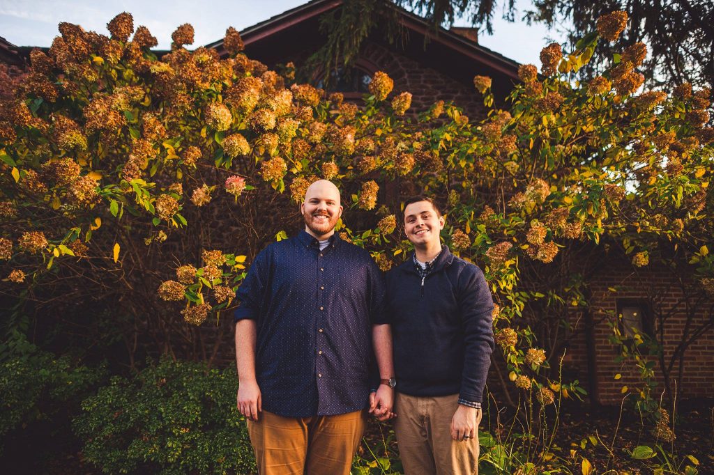 Kevin and Scott stand holding hands in front of golden hydrangeas out front of the Hollybush Mansion. 