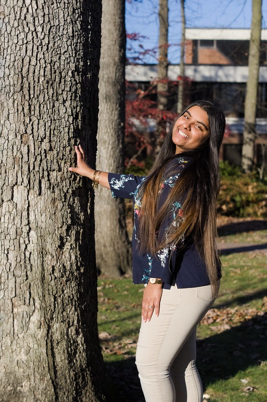 Riya stands beside a tree, smiling.