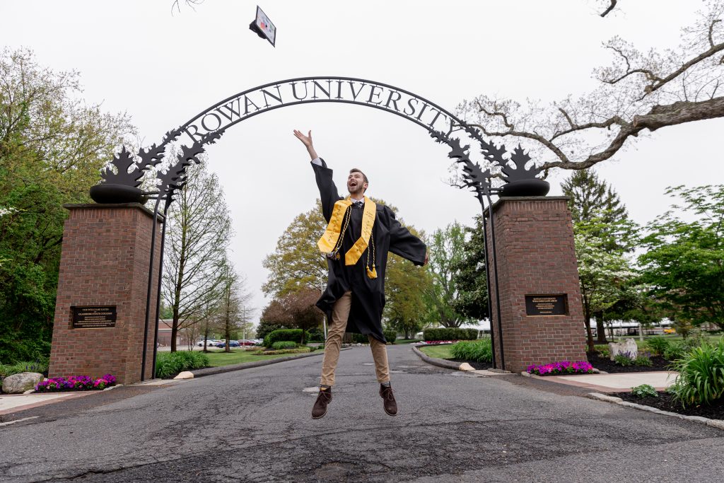 Griffin jumps and throws his graduation cap in the air in front of the Rowan arch.