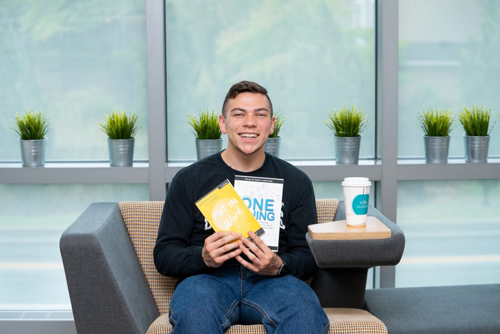 Josh sits holding two business textbooks and smiling inside Business Hall.