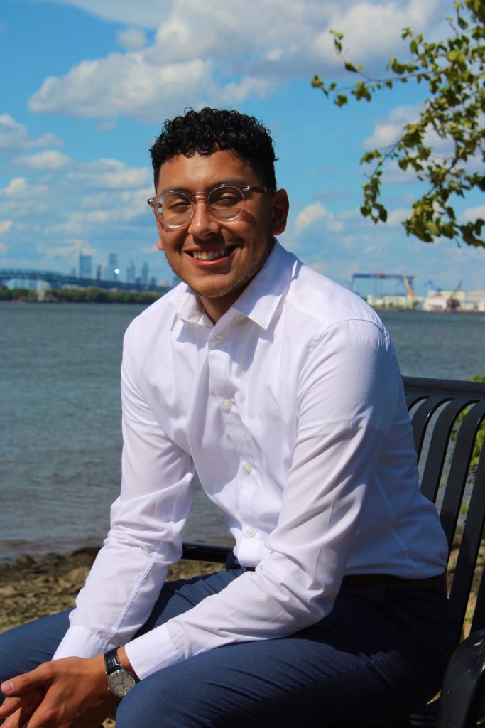 Jacob sits on a bench beside a River with the city in the background.