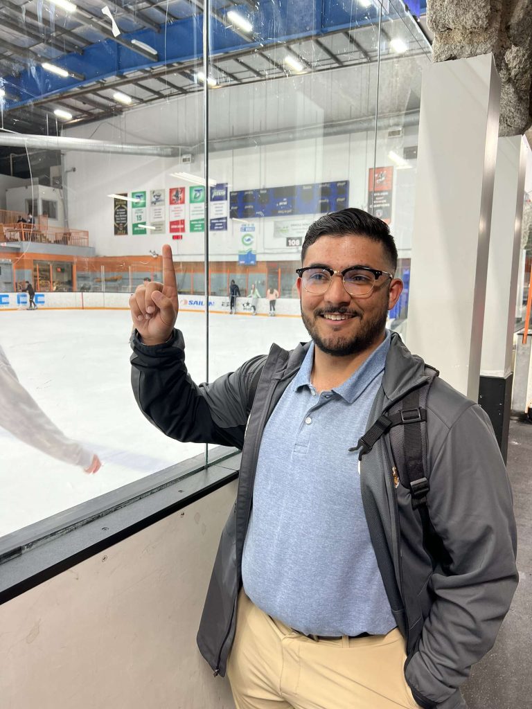 Spencer Reyes pointing up at the scoreboard, standing next to an ice hockey rink.
