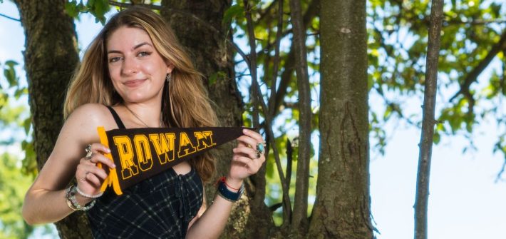 Skyla holds a Rowan University pennant against a wooded backdrop.