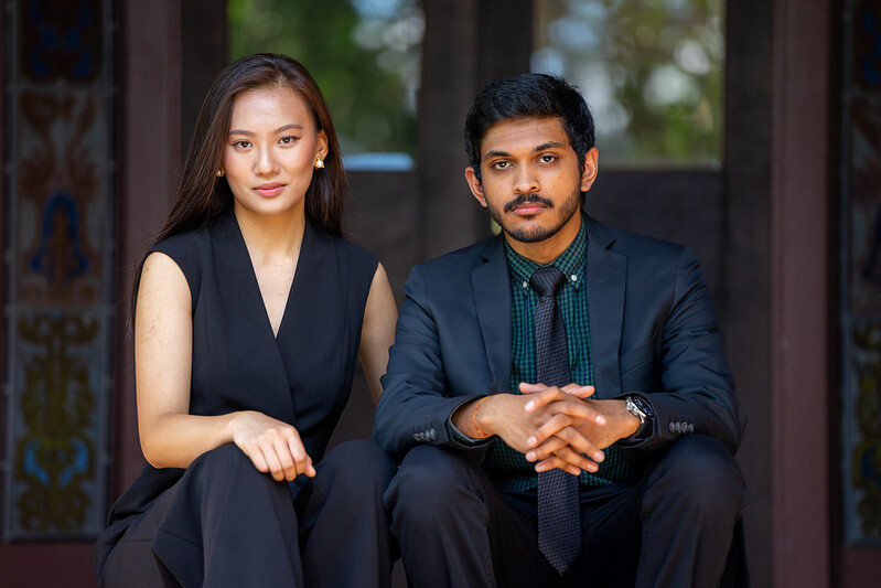 Naman and Terry are sternly looking directly into the camera while sitting next to each other. 