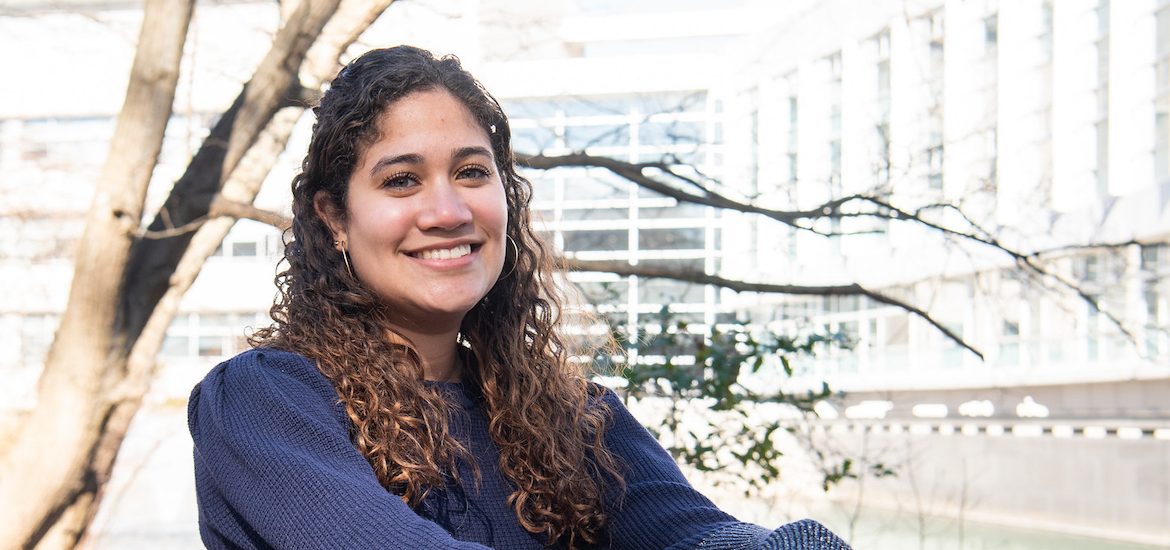 Maria is sitting and smiling with Rowan Hall in the background.
