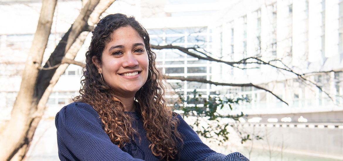 Maria is sitting and smiling with Rowan Hall in the background.