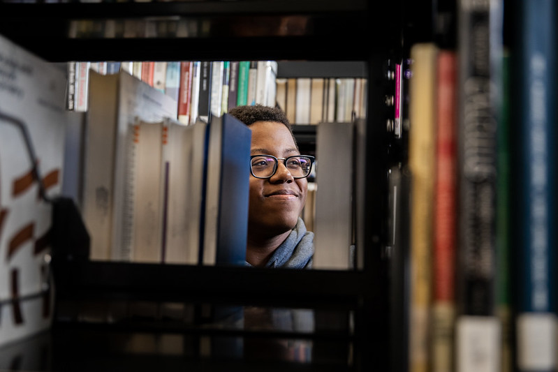 Rowan Health and Science Communication major Sedrick smiles between book stacks inside Campbell Library.