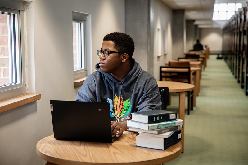 Rowan Health and Science Communication major Sedrick works on a laptop and stares out the window of Campbell Library.