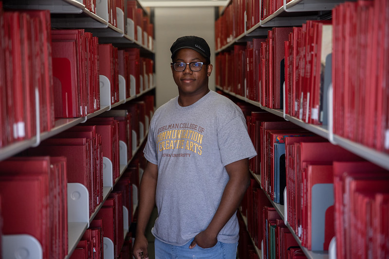 Rowan University Health and Science Communication major Sedrick stands inside the stacks in between two bookshelves on campus.