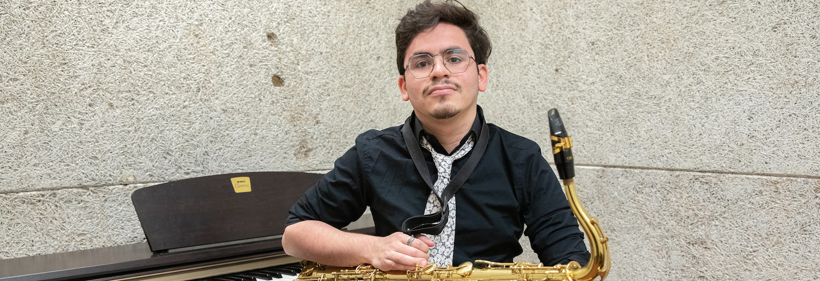 Rowan University student Jovan Rivera posing inside of Wilson Hall with a saxophone sitting in front of a piano.
