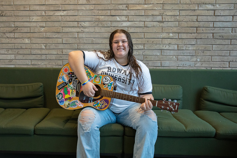 Laynie sits inside Discovery Hall on a green couch holding a guitar covered in decorative stickers.