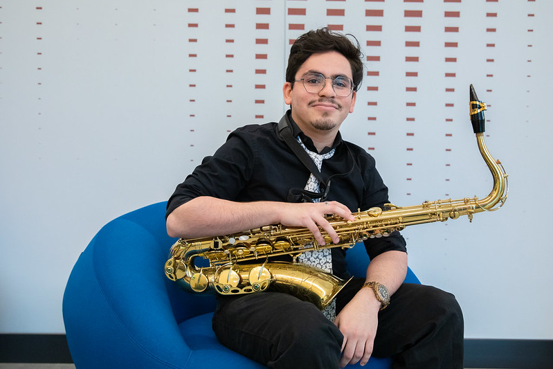 Rowan University student Jovan Rivera smiles and holds a saxophone while sitting in a bright blue chair. 