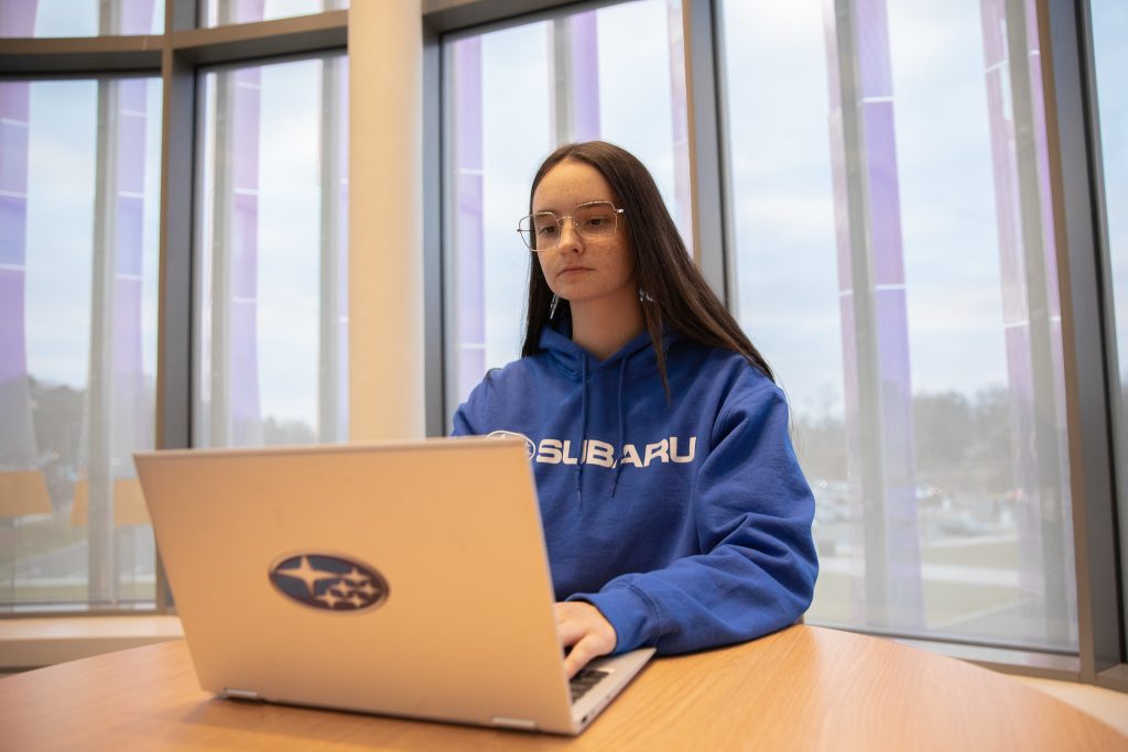 Rowan University Accounting major Jade Kenny wears a blue sweatshirt and works on a laptop with the Subaru logo in Business Hall.