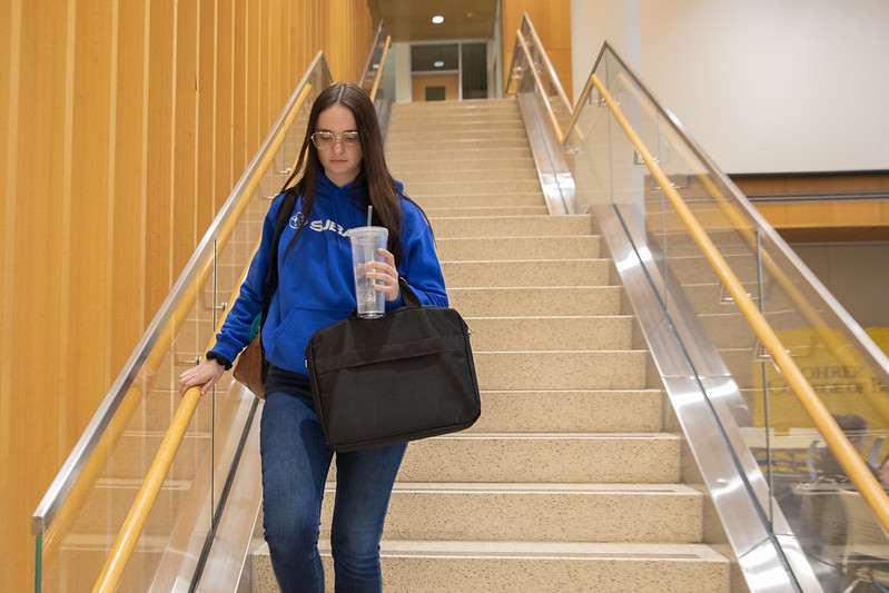 Rowan University Accounting major Jade Kenny wears a blue sweatshirt and walks down the stairs in the lobby of Business Hall.