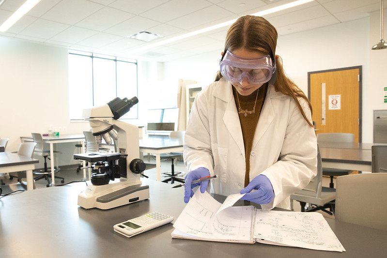 Rowan University Biological Sciences major Mia Shute looks over her notes in a lab wearing protective goggles and a lab coat.