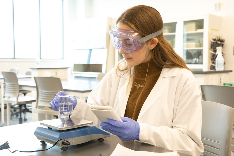 Rowan University Biological Sciences major Mia Shute works on an experiment in a lab wearing protective goggles and a lab coat.
