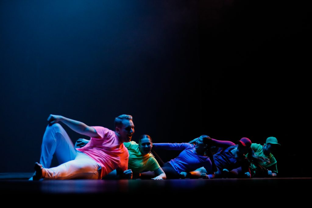 Five dancers pose while on the floor, with rainbow colored lights illuminating them. 
