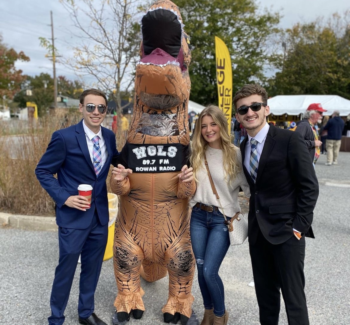 Danny Ryan wears business attire while posing with the Rowan Radio dinosaur and two friends at Rowan Homecoming.